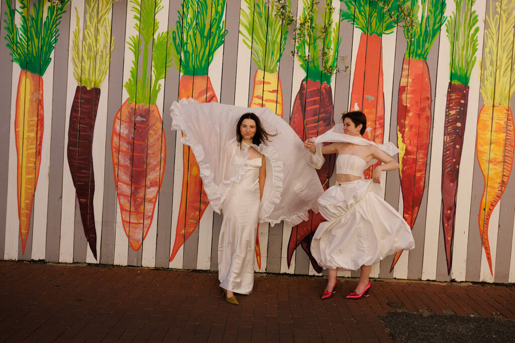 Two brides pose for wedding portraits in front of a carrot mural in Washington DC. One bride is queer wedding stylist, Kati Kons from Portrait of a Bride on Fire.
